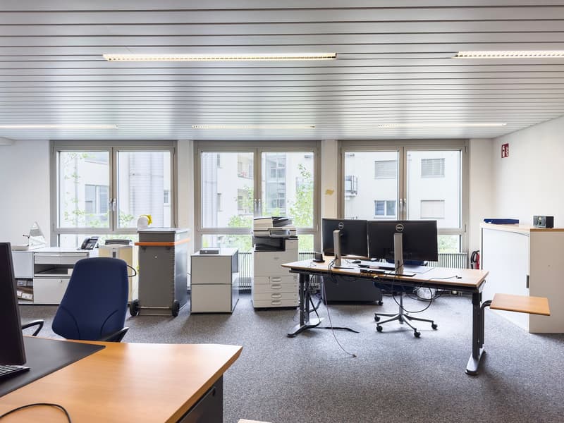 The image shows an office space with multiple workstations. The room has large windows providing natural light, and is equipped with various office equipment such as computers, printers, and filing cabinets. The space appears to be well-organized and desi
