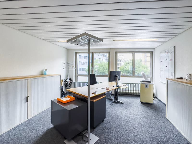 The image shows a modern office space with large windows, a wooden desk, a computer setup, and various office equipment and furniture. The room has a minimalist and clean design with white walls, a gray carpet floor, and a suspended ceiling with linear li
