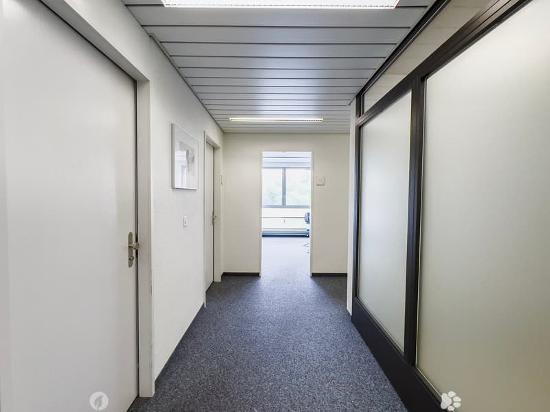 The image shows a long, narrow hallway with a gray terrazzo floor and white walls. The ceiling has a series of parallel beams running across it, creating a modern, industrial look. There are several doors lining the hallway, suggesting this is an office o