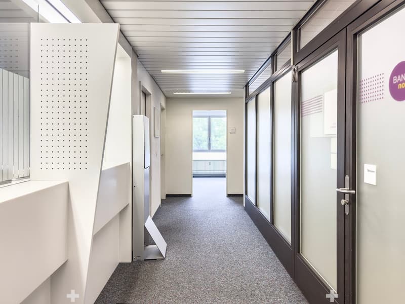 The image shows a long, well-lit hallway in an office or commercial building. The hallway has a gray terrazzo floor, white walls, and a suspended ceiling with fluorescent lighting. There are several doors and glass panels along the hallway, as well as som