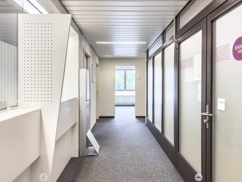 The image shows a long, well-lit hallway in an office or commercial building. The hallway has a gray terrazzo floor, white walls, and a suspended ceiling with fluorescent lighting. There are several doors and glass panels along the hallway, as well as som