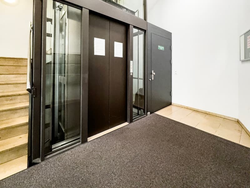 The image shows the entrance to a building. The entrance has a set of double glass doors with a black frame. The doors are flanked by two solid black doors on either side. The floor in front of the entrance is covered with a gray carpet. There are wooden
