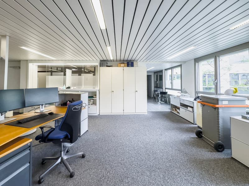 The image shows a modern office space with a gray and yellow color scheme. The room has a high ceiling with a paneled design, and large windows that provide natural light. There are multiple workstations with computers and office chairs, as well as storag