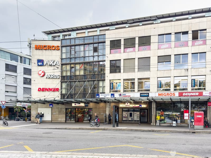 Multi-story commercial building with various retail stores and businesses, including Migros, Axa, Akros, and Denner. The building has a modern glass and concrete facade with signage for the different companies. There are parking spaces in front of the bui