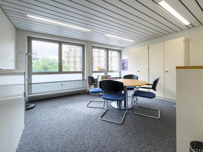 The image shows a modern office space with a large window overlooking the city. The room has a gray carpeted floor, a round wooden table surrounded by black chairs, and a wall-mounted storage unit. The ceiling is lined with gray metal panels and features