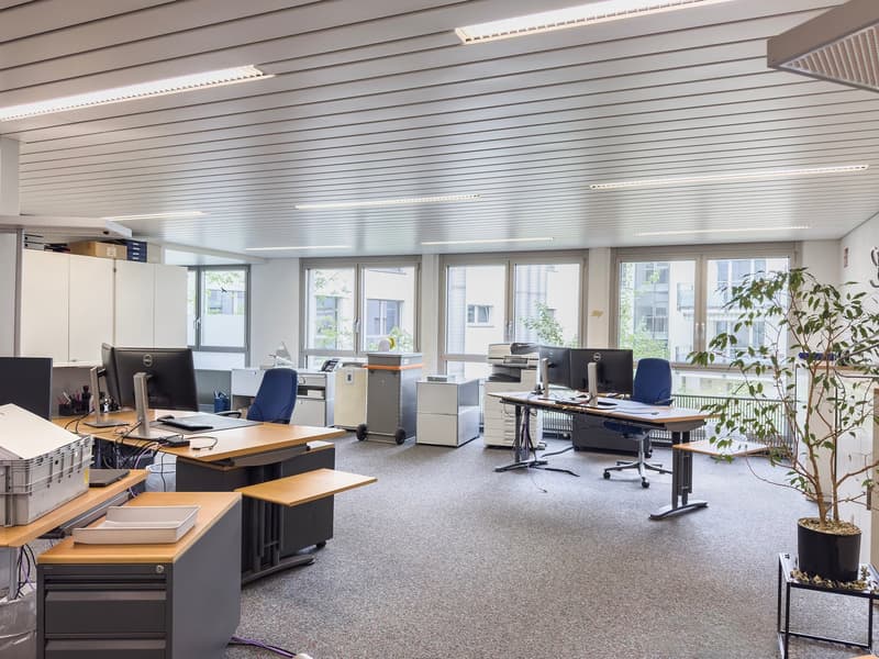 The image shows a modern office space with large windows, a suspended ceiling with lighting fixtures, and various office furniture and equipment such as desks, computers, printers, and a plant. The space appears to be well-lit and organized, with a profes