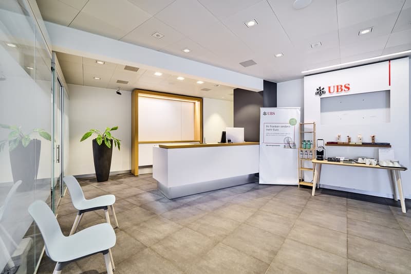 Modern office reception area with white desks, wooden shelves, white chairs, large potted plants, and UBS branding.