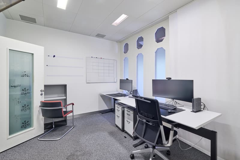 minimalistic office room, gray carpet, 2 desks with chairs, computer monitors, white walls, small bookshelf, glass door