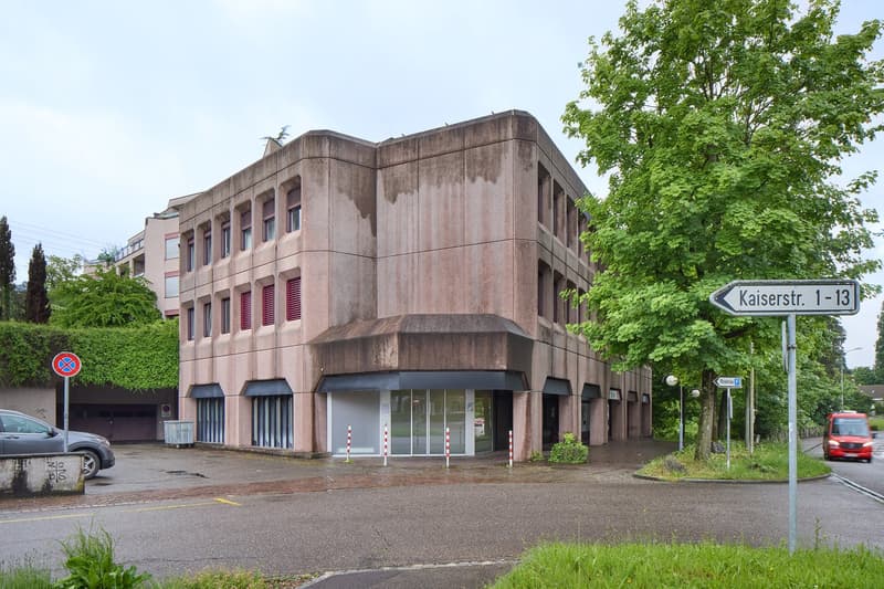Large building with multiple floors, large windows, small parking in front.