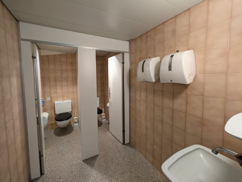 Bathroom with beige tiles, white fixtures, two toilets, and two hand dryers.