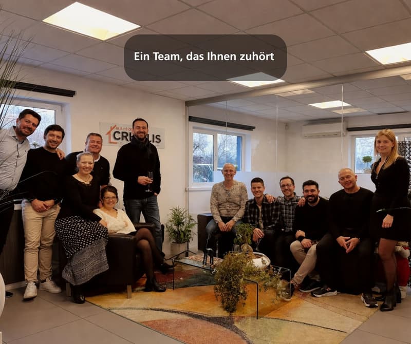 group of people standing and sitting in a room, with a carpet, plants, and a glass table