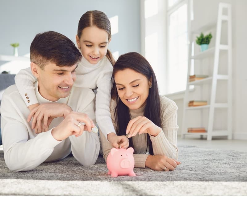 family putting coins in piggy bank, white walls, carpet floor, shelves with plants and books