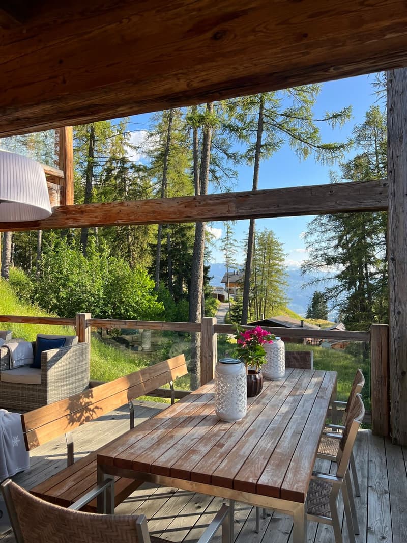 Wooden patio with a dining table and chairs, surrounded by a lush forest landscape with tall pine trees. The patio is covered by a wooden roof structure, providing a sheltered outdoor living space with a scenic view.
