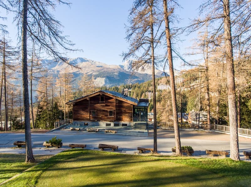 Wooden cabin-style house in a forested mountain setting, with a balcony or terrace overlooking the surrounding landscape. The house is surrounded by pine trees and has a grassy area in front.