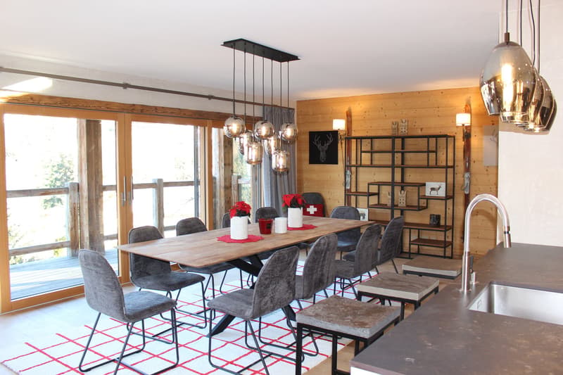 Spacious dining area with a large wooden table and grey upholstered chairs. The room features a modern lighting fixture with glass pendants, and a wooden accent wall. Large windows provide a view of the outdoor area. The room also includes a metal shelvin