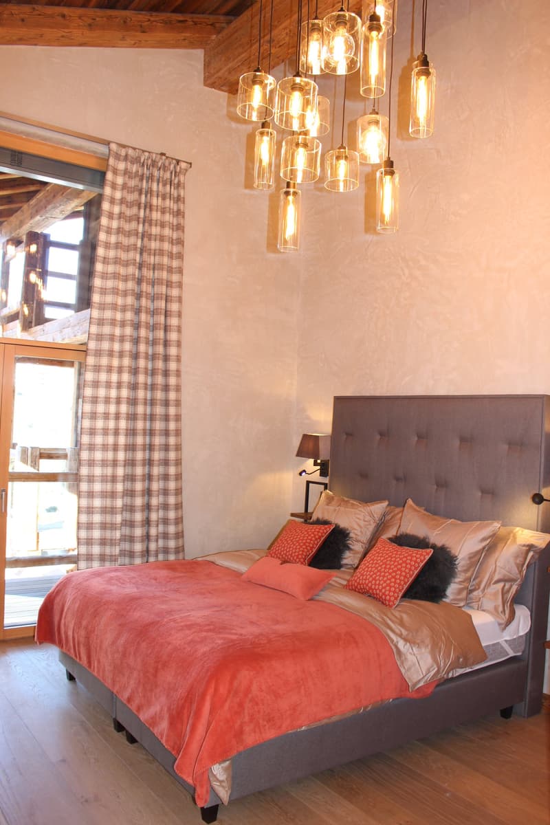 Cozy bedroom with wooden ceiling, large windows, tufted headboard, orange and gold bedding, and a cluster of pendant lights above the bed.