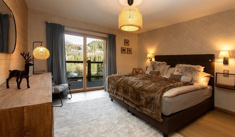 Spacious bedroom with large windows overlooking a scenic mountain view, a plush king-size bed with a fur blanket, wooden furniture, and decorative lighting fixtures.