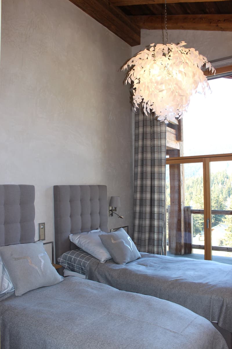 Spacious bedroom with gray upholstered headboard, plaid curtains, and a large feather chandelier. The room has a cozy and rustic feel with the exposed wooden beams on the ceiling.