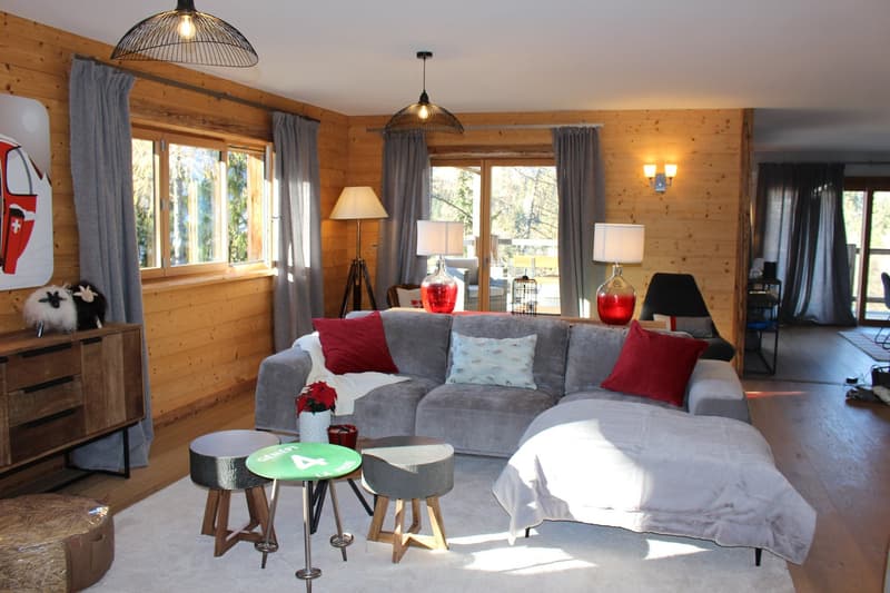 Cozy living room with wooden walls, large windows, gray sofa with red and patterned pillows, coffee table with stools, floor lamps, and a balcony visible through the windows.