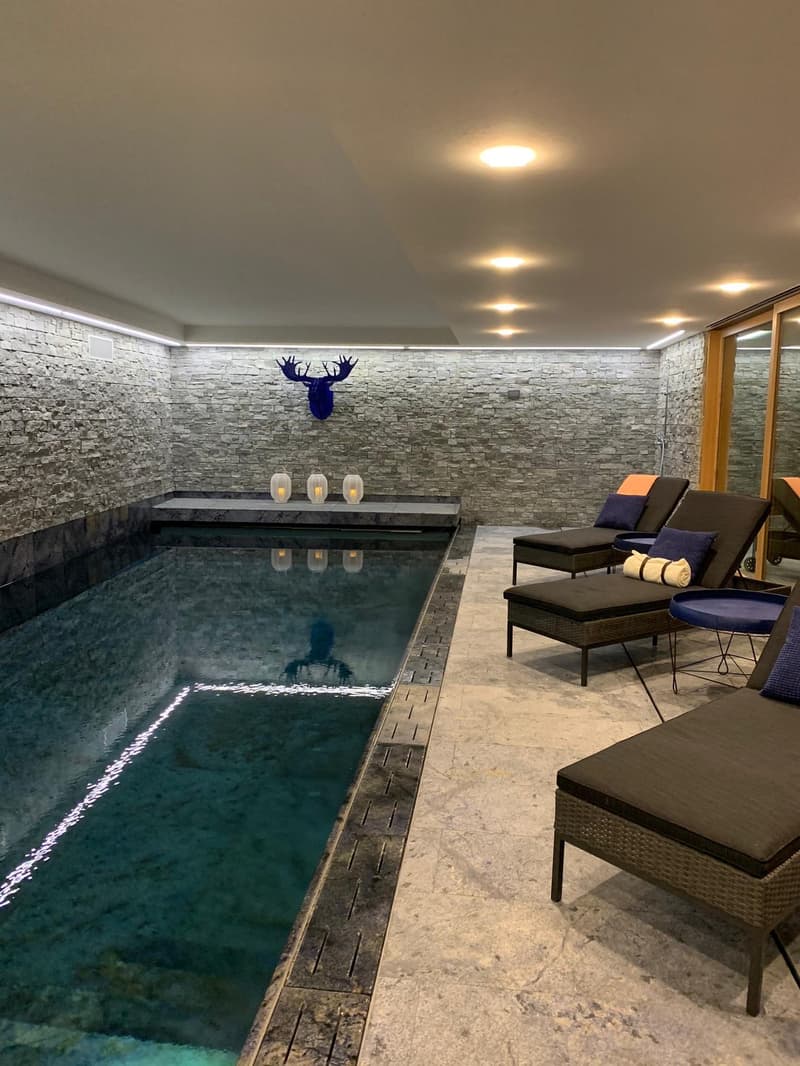 The image shows an indoor pool area with a rectangular pool surrounded by a stone wall. The pool has a dark blue color and there is a reflection of the ceiling visible in the water. The room has a modern and cozy design, with comfortable lounge chairs and