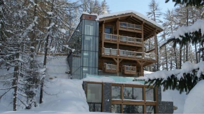 2-story wooden chalet-style house with large windows, balconies, and a glass entryway, surrounded by snowy pine trees