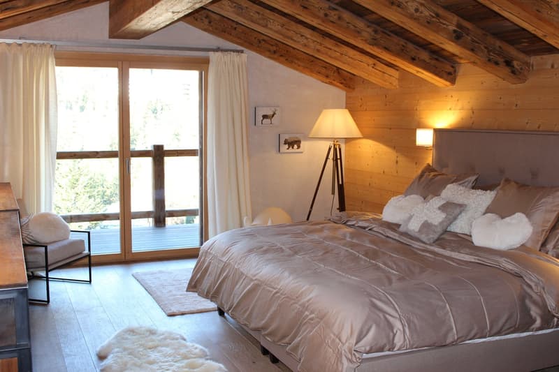 Cozy bedroom with wooden beamed ceiling, large windows with balcony access, plush bedding, and rustic decor elements like the animal artwork and tripod lamp