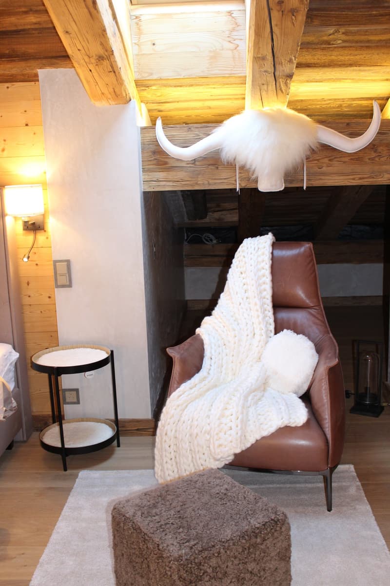 Cozy living room with wooden walls and ceiling, a leather armchair with a knitted blanket, a side table, and a decorative animal horn mounted on the wall.