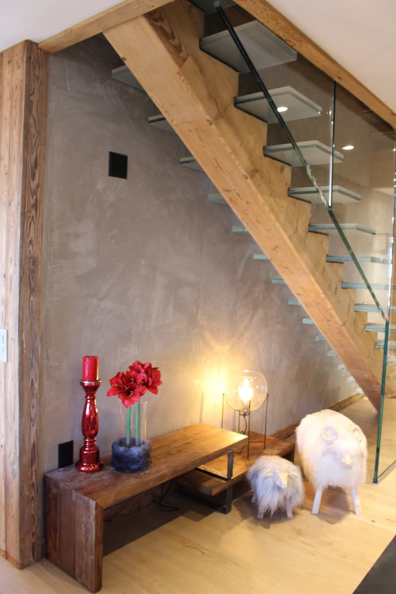 The image shows a cozy interior space with a wooden staircase, a wooden console table with decorative items like a red candle, flowers, and a lamp, and a fluffy white chair. The walls have a textured, gray finish and the floor appears to be hardwood. The