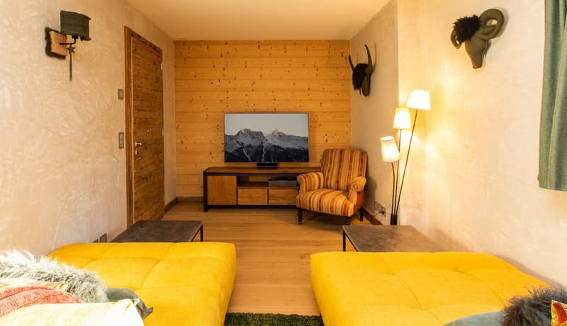 Cozy living room with wooden walls, a large TV mounted on the wall, a yellow sofa, a coffee table, and a fireplace. The room also features decorative elements like animal horns on the wall and lamps.