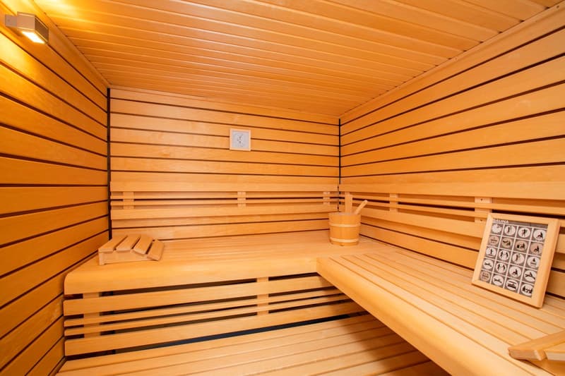 The image shows the interior of a wooden sauna room. The walls and ceiling are made of wooden panels, and there are wooden benches along the walls. A sauna sign is visible on the wall, indicating this is a sauna room. The room appears to be well-lit with