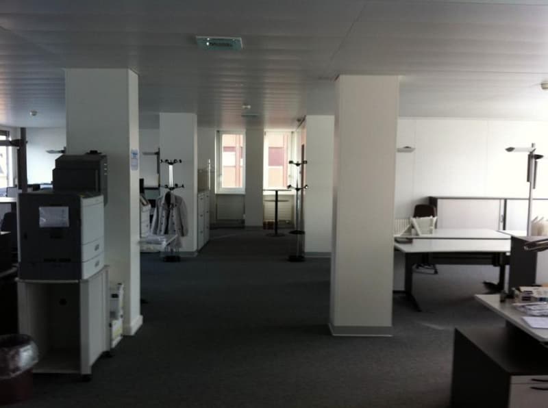 Large open office space with white walls, black carpeting, and various office equipment such as printers, computers, and a refrigerator. There appears to be a private washing machine and dishwasher visible in the room.