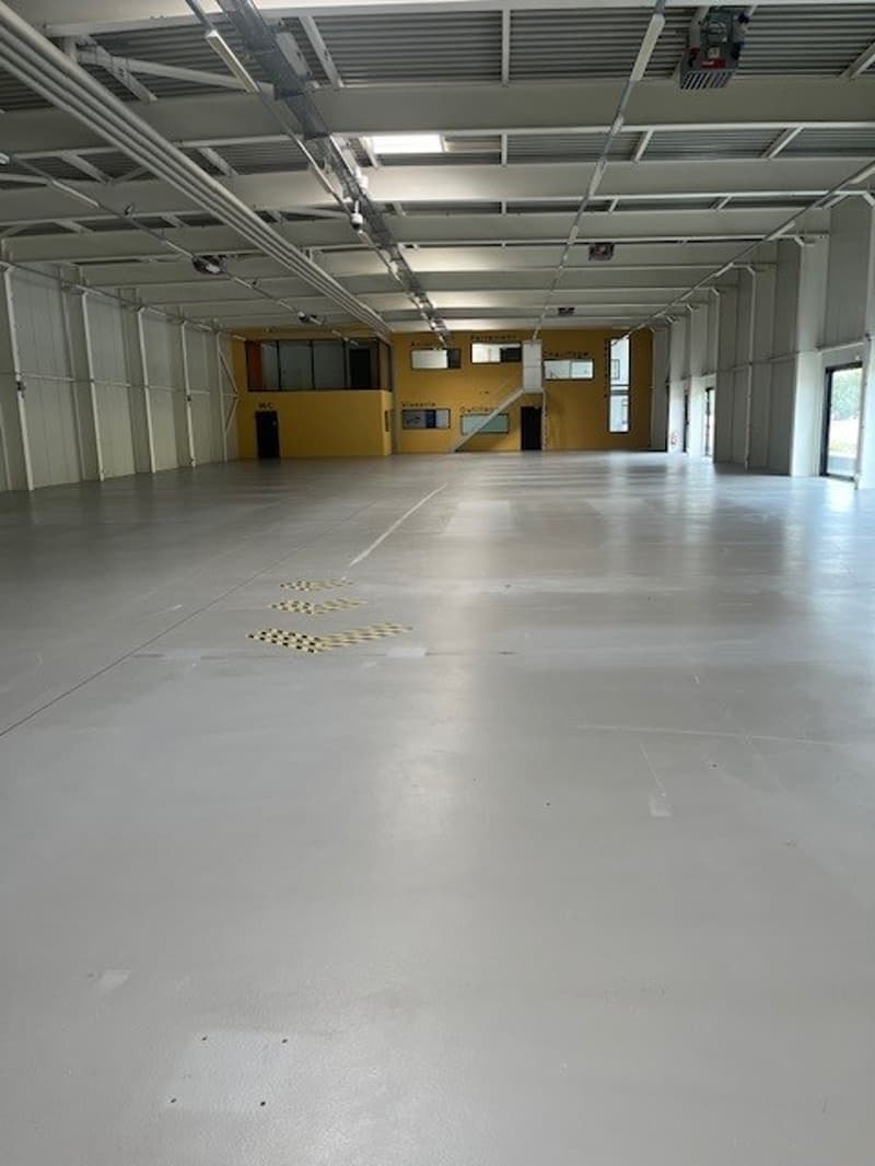 Large open commercial space with concrete flooring, exposed ceiling beams, and several windows. The space appears to be empty and unfinished, likely a warehouse or industrial facility.