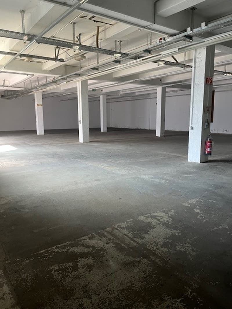Large open space with concrete floor, white walls, and overhead lighting fixtures. The space appears to be a warehouse or storage facility, with a visible garage door or entrance.