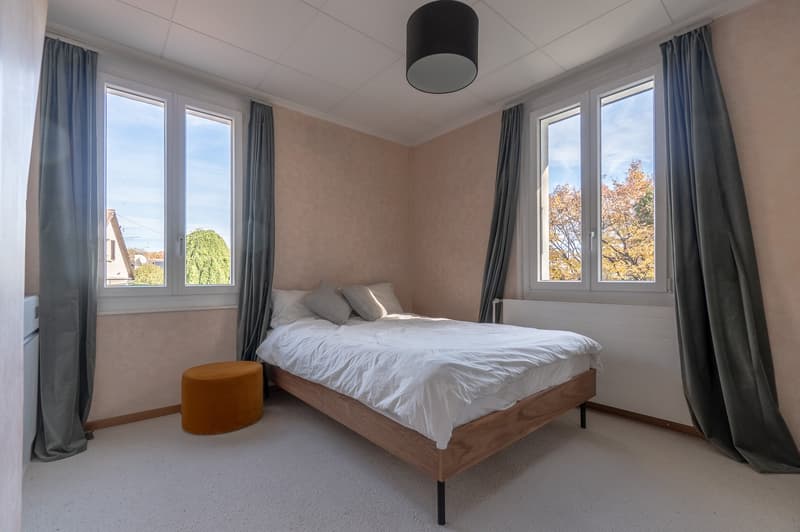 Bedroom with double bed, 2 pillows, gray curtains, white walls, two large windows, brown round ottoman