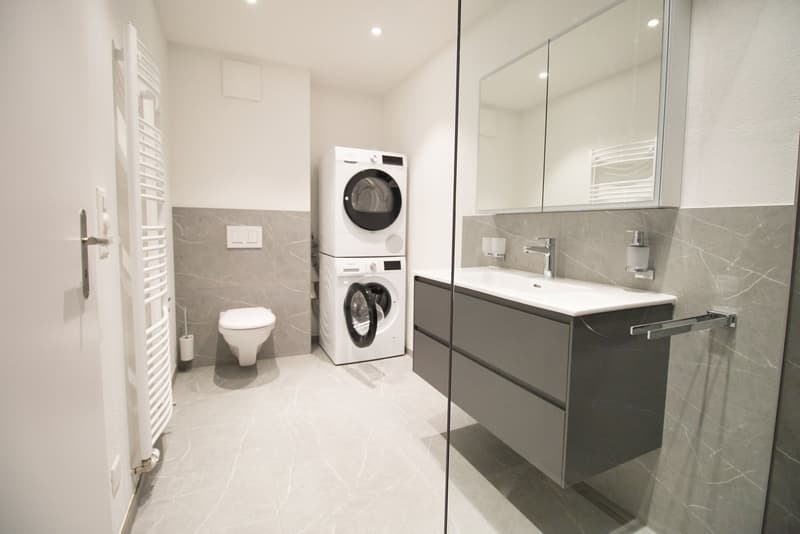 modern bathroom with white tiles, white toilet, white washing machine, white sink, mirror, and towel rack