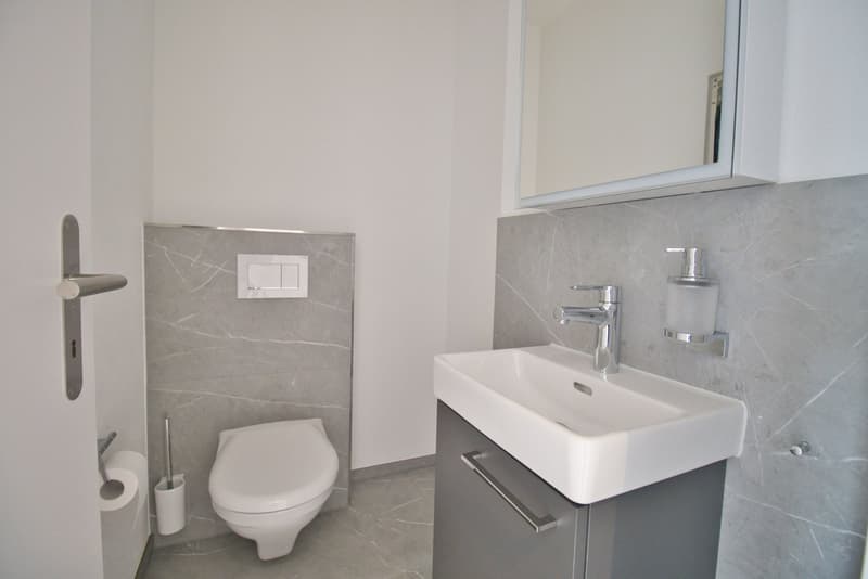 modern bathroom with white toilet, sink, mirror, and gray tiles