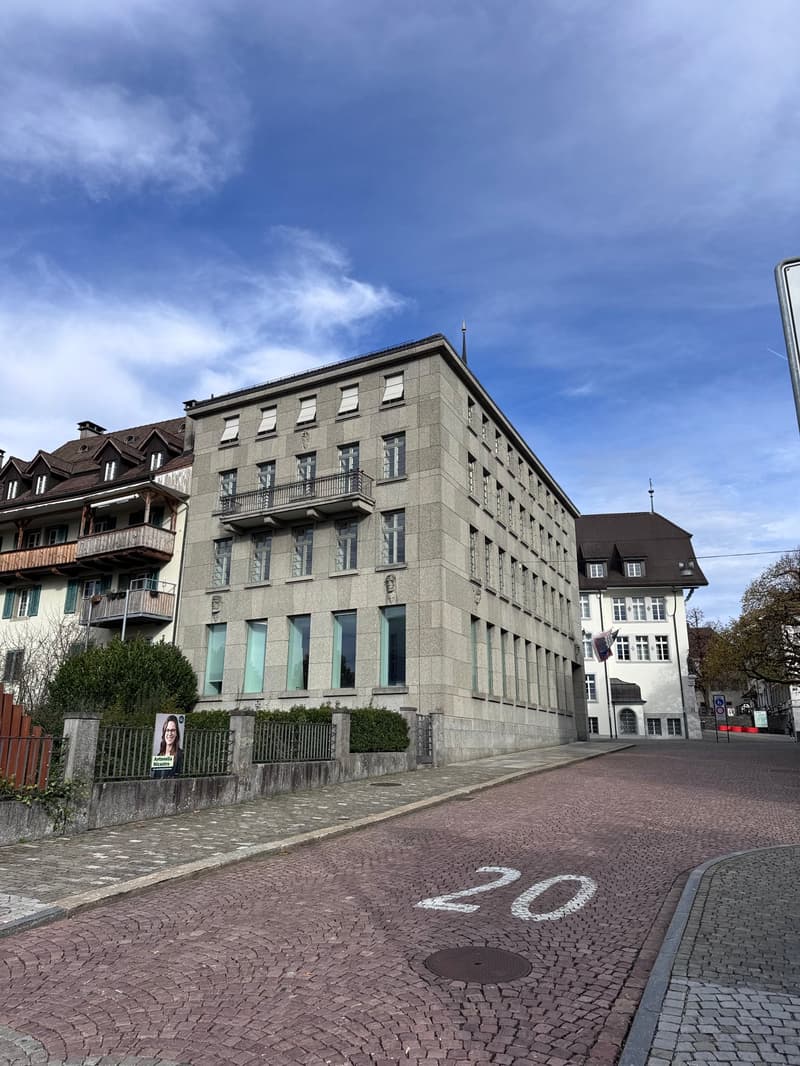 4 story residential building with multiple balconies, brown roof, large windows, flag on the building