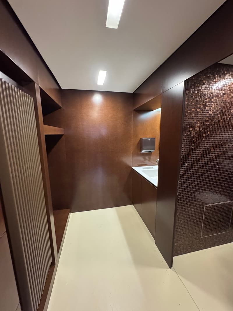 The interior of a room features walls and floors covered in brown materials. There is a white sink with faucet, a wooden cabinet, and a tissue paper dispenser on the wall.