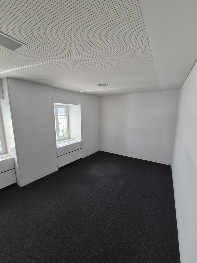 Empty room with black carpet, white walls, windows, built-in bench with drawers