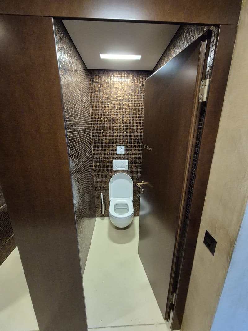 Compact toilet space, white toilet bowl, brown walls, white flooring, built-in lighting, open door
