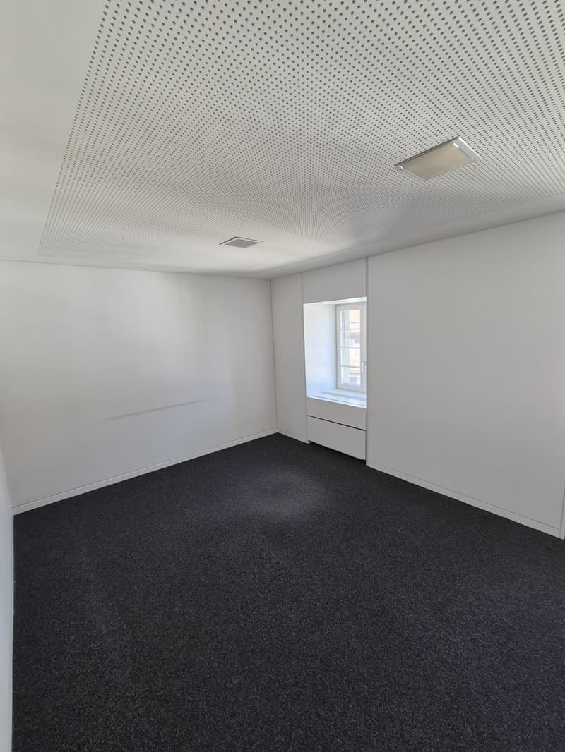 An empty room with black carpet flooring, white walls, a small window, and a ceiling with a square pattern and a built-in light.
