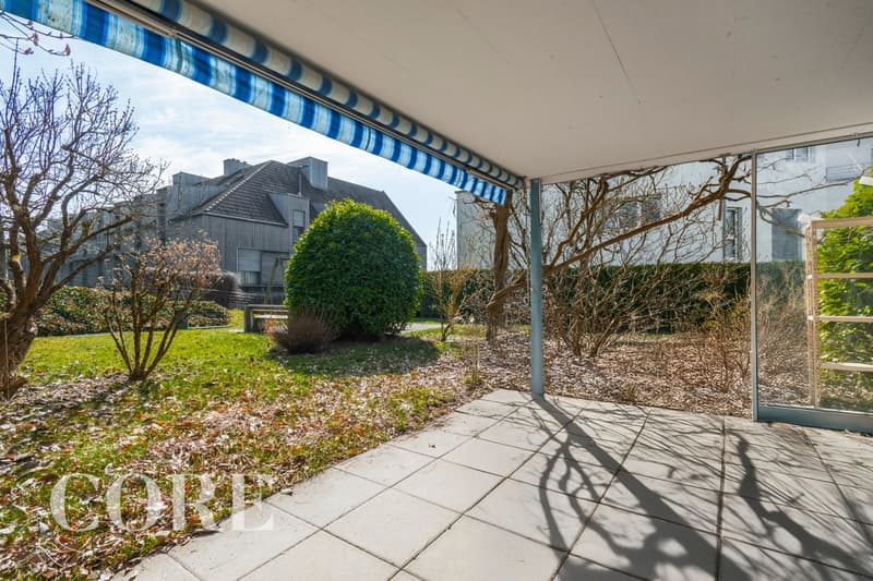 covered terrace with blue and white striped awning, tiled floor, glass door, garden with shrubs and trees, pathway, and distant buildings