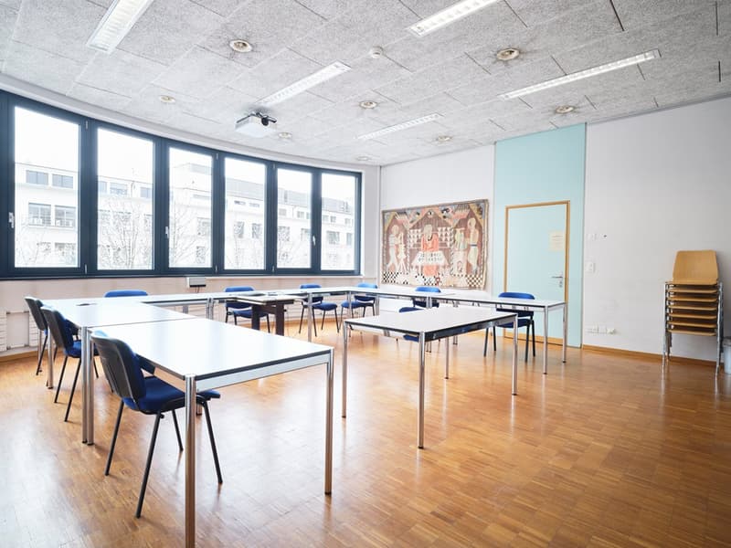 classroom with wooden floor, white desks, blue chairs, large windows, projector, radiator, blue and white wall, artwork