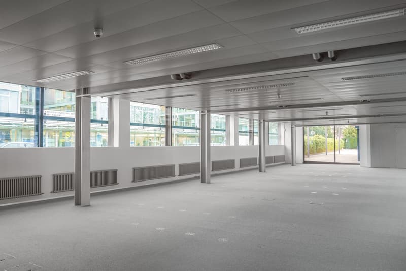 An empty office space with a ceiling featuring recessed lighting, glass windows, central heating radiators, and a glass door leading to the exterior. No visible furniture or personal belongings.