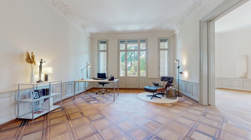 Large, well-lit room with parquet floor, desk, office chair, and cozy lounge chair with a table. There are several windows providing natural light and views of the outside. A white shelf holds books and a decorative vase. Two standing lamps on either side