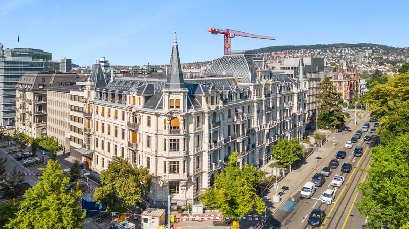 Large, multi-story building with multiple spires and turrets, surrounded by greenery and city buildings, with a construction crane on top of one of the nearby buildings