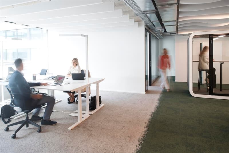 Two work stations with laptops, glass-walled cubicle, white and green carpeted floors, people