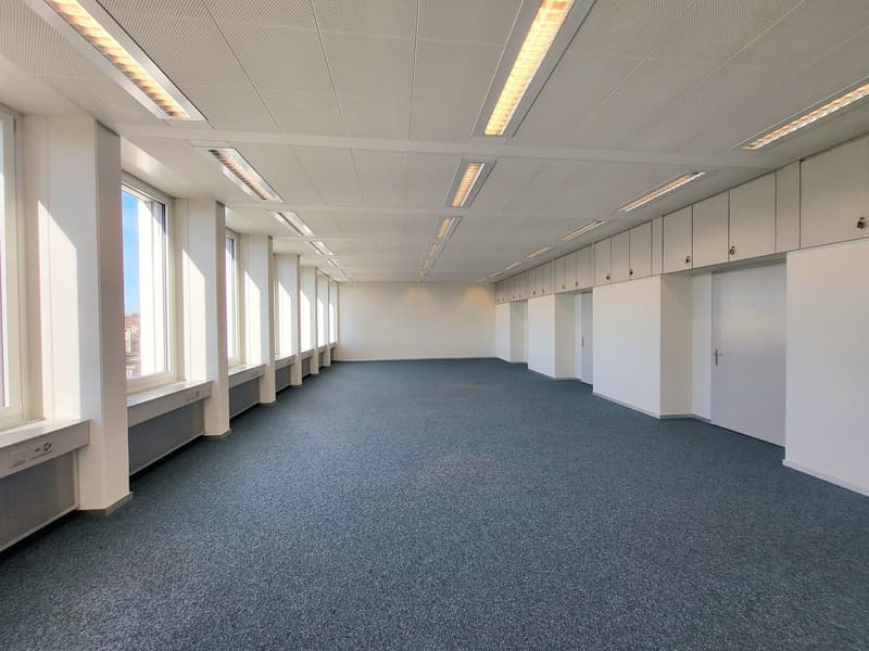 Office space with gray carpeting, multiple large windows, white walls, white ceiling with grid lighting, and a door on the far right side.