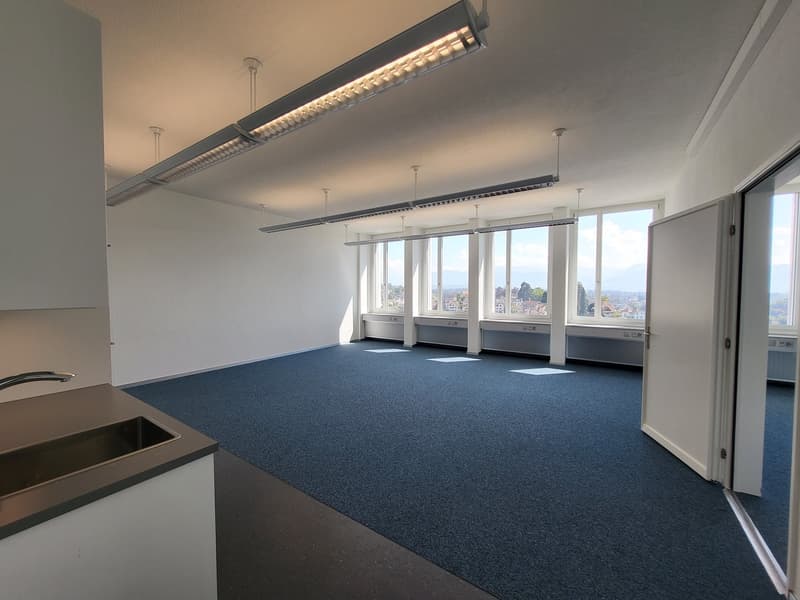 Large open office space with floor-to-ceiling windows providing a panoramic view of the city. The room has a blue carpeted floor, recessed lighting fixtures, and a kitchenette area.