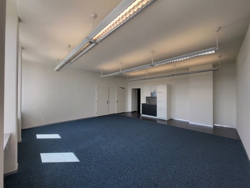 The image shows a large, open office space with a gray and blue color scheme. The room has a high ceiling with recessed lighting fixtures and a long, rectangular light fixture running along the ceiling. The floor is covered in a dark blue carpet, and ther